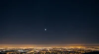 Céu noturno estrelado com planeta visível a olho nu, cidade iluminada ao fundo e cores azuis, pretos, laranjas e amarelos, representando descubra universo como avistar
