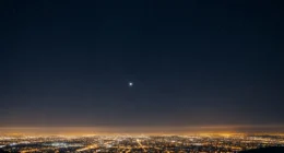 Céu noturno estrelado com planeta visível a olho nu, cidade iluminada ao fundo e cores azuis, pretos, laranjas e amarelos, representando descubra universo como avistar