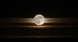Imagem da Lua no céu noturno parcialmente coberta por nuvens finas, com a Terra no canto inferior esquerdo, mostrando o mistério da lua que vemos